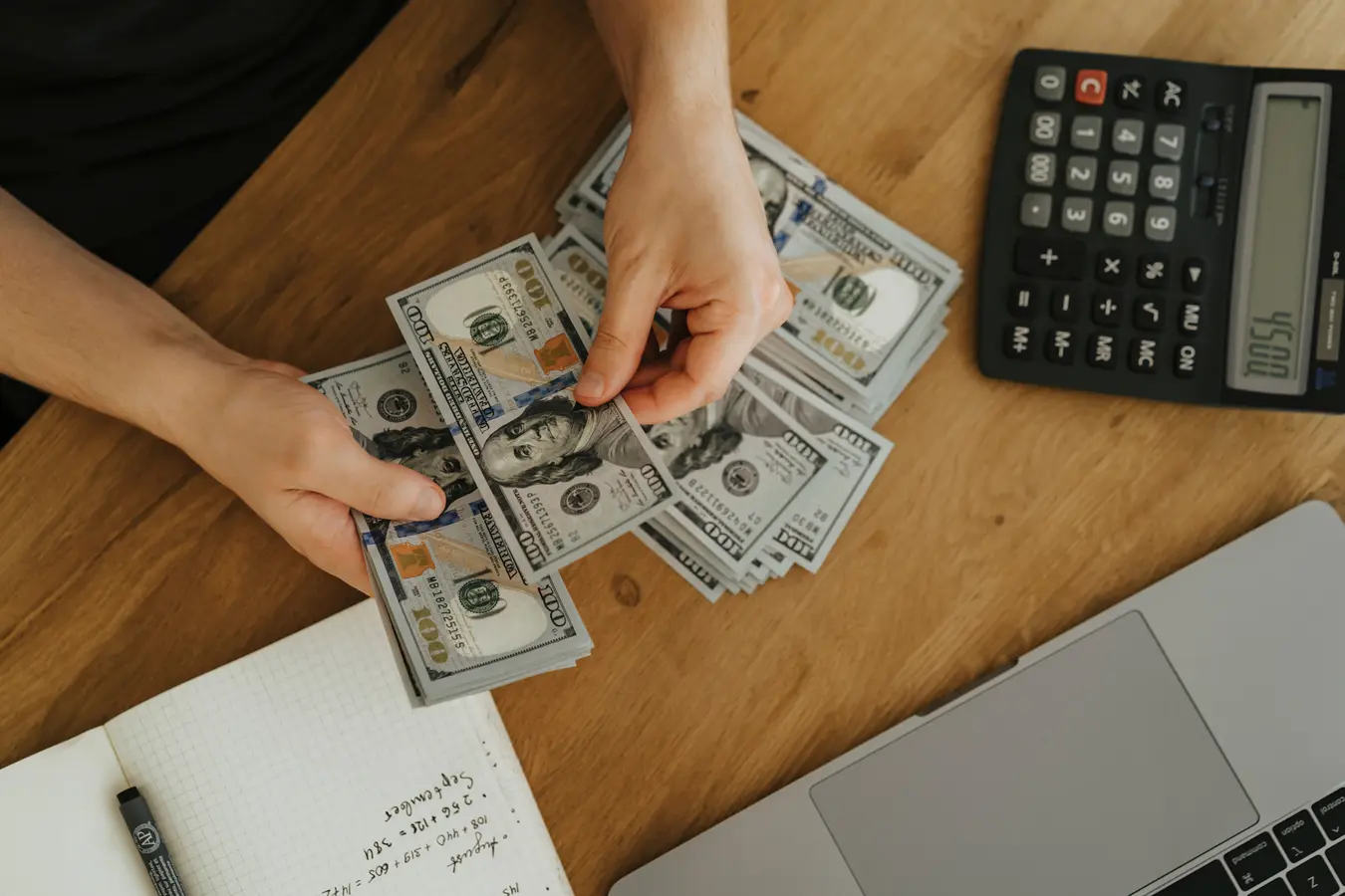 Hands counting dollar bills beside a calculator, notebook, and laptop for mortgage payment planning