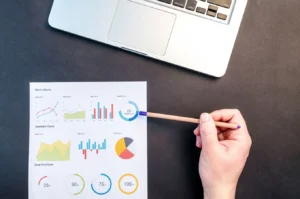 Hand holding pencil reviewing colorful data charts on desk beside laptop for Google intrinsic value analysis