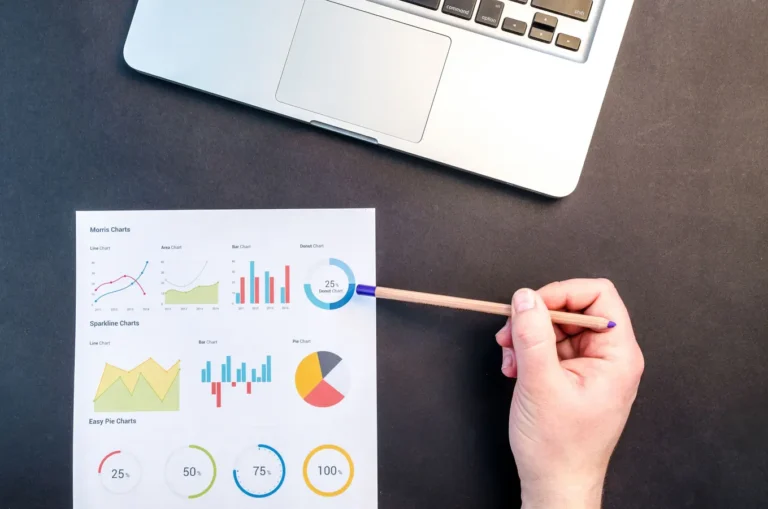 Hand holding pencil reviewing colorful data charts on desk beside laptop for Google intrinsic value analysis