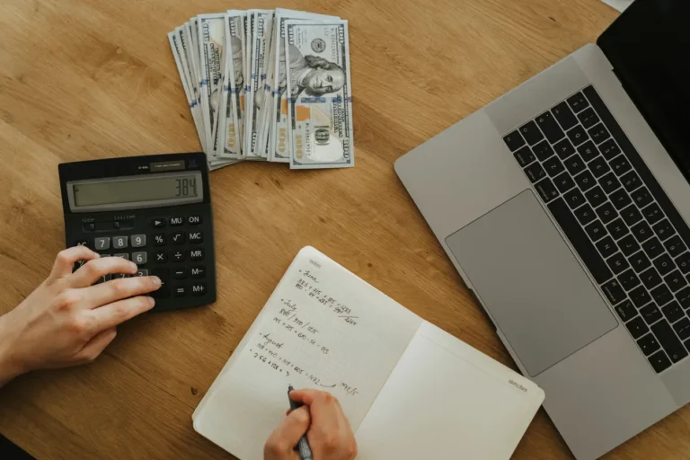 Hands using a calculator and notebook to plan mortgage loan EMI with cash and laptop