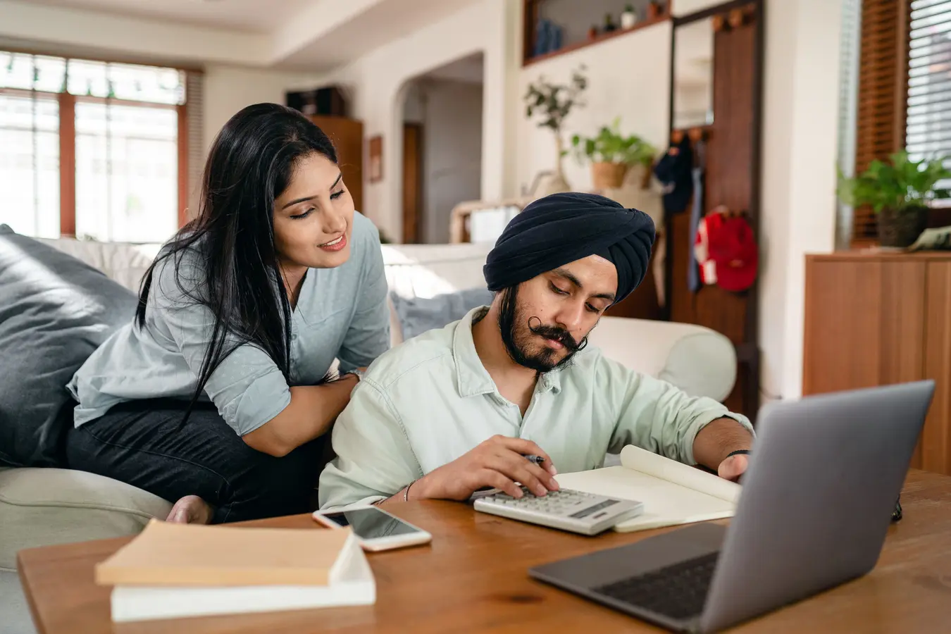 Woman on sofa watching husband calculate mortgage loan EMI at coffee table with laptop and calculator