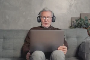 Senior man with headphones using a laptop on a gray couch at home