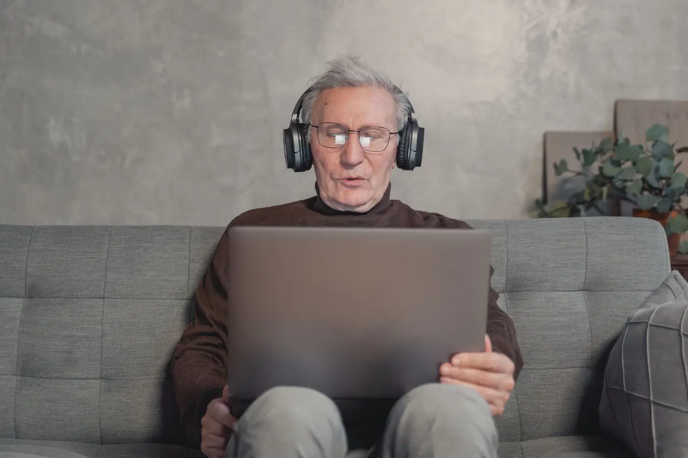 Senior man with headphones using a laptop on a gray couch at home