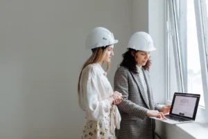 Two women in hard hats review construction plans on a laptop, reflecting Procore project management software use