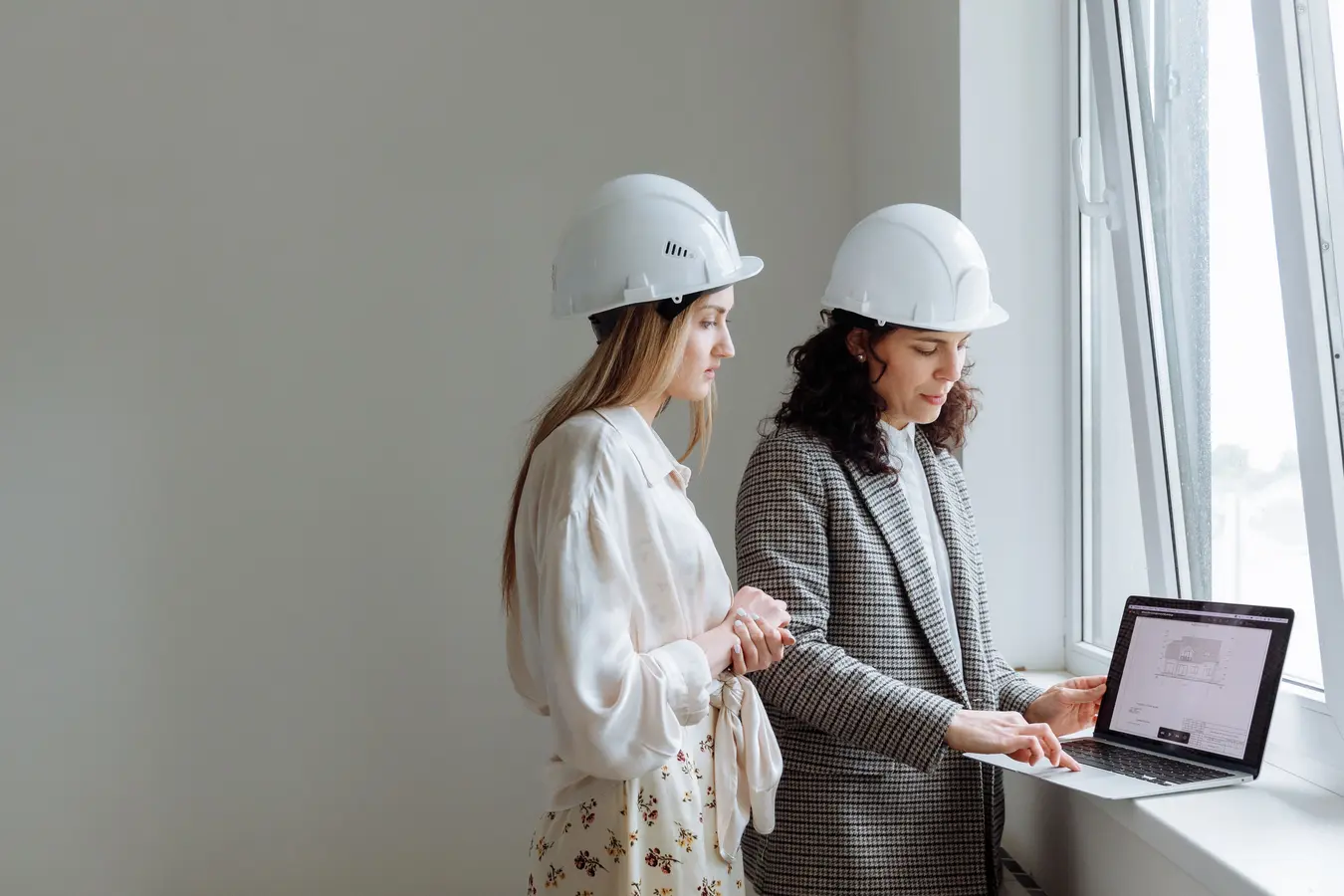 Two women in hard hats review construction plans on a laptop, reflecting Procore project management software use