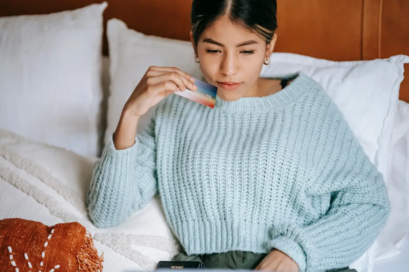 Woman lying on cozy bed in blue sweater holding a credit card